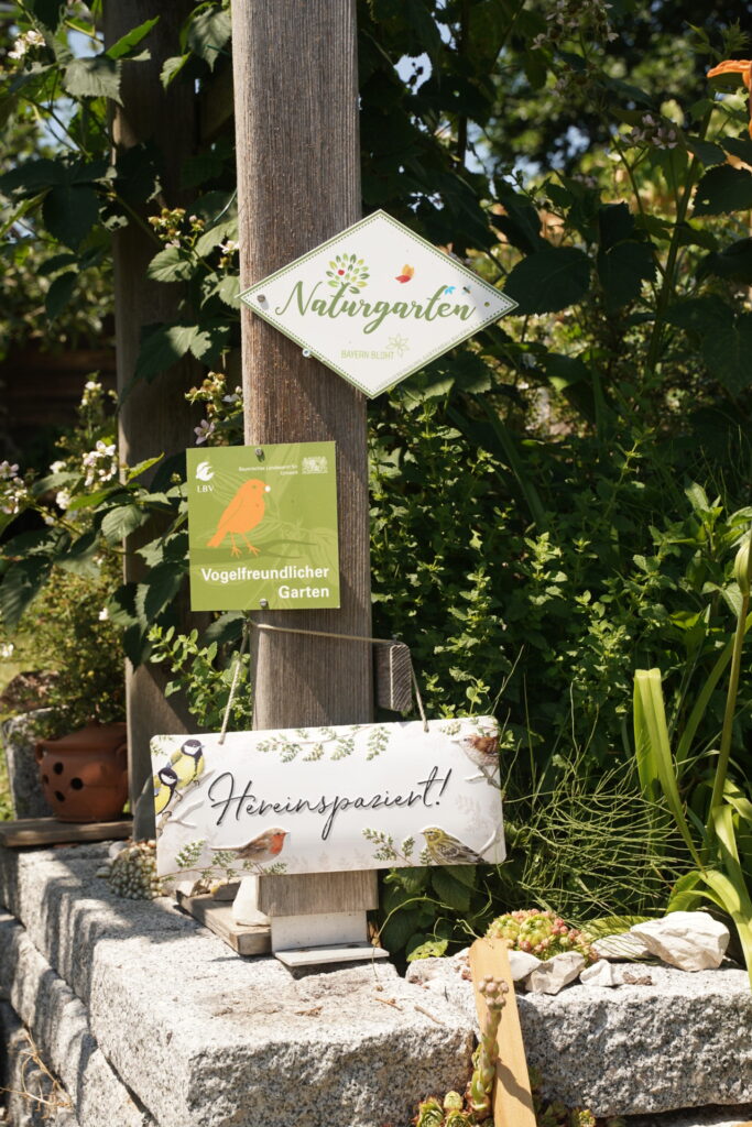 An einem Holzpfosten sind drei Plaketten angebracht, auf denen steht: "Vogelfreundlicher Garten", "Naturgarten" sowie "Hereinspaziert"